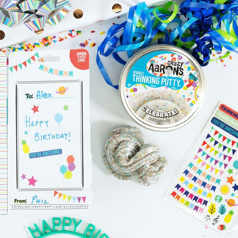 Tin of Celebrate Thinking Putty® on a table with a greeting card, ribbons and sheet of stickers.