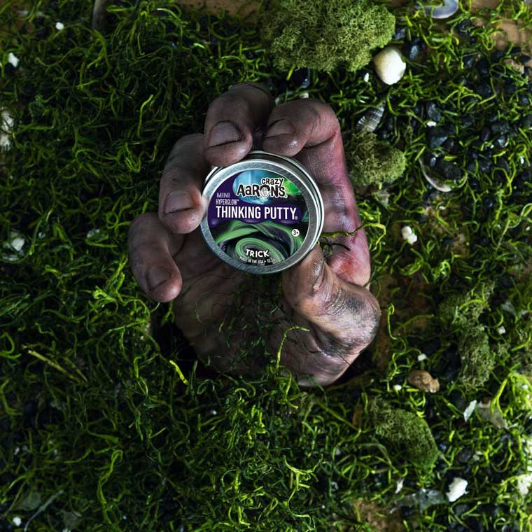 Hand coming out of a grass and dirt scene holding a mini tin of Trick Thinking Putty®.